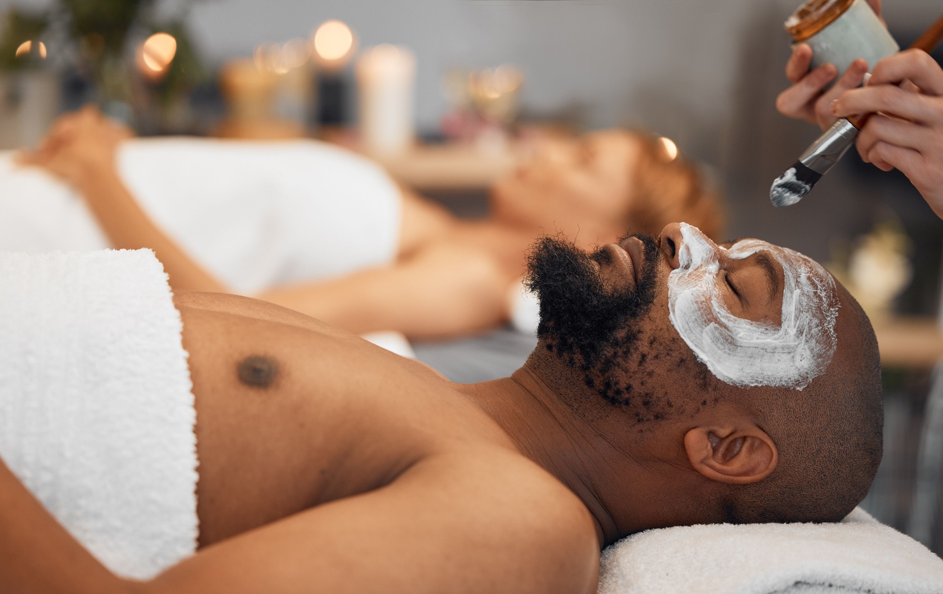 Skincare, wellness and black man getting a facial at a spa for relaxation, calm and satisfaction. Beauty, luxury treatment and couple massage at beauty salon together, skincare products on man's face